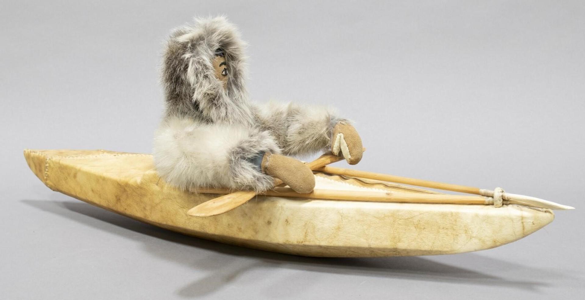 Inuit Model Kayak With Paddler - a sealskin covered wood-framed kayak containing a figure wearing a fur parka, deerskin gloves and holding a wooden paddle; with mounted wood, bone and sinew spears
